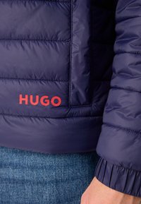Mørkeblå pufferjakke med quiltet tekstur, elastiske mansjetter og rødt "HUGO"-logo nederst til venstre. Materialet ser glatt ut og er vannavvisende.