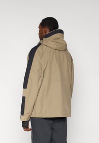 Μπεζ και ναυτικό μπουφάν με κουκούλα, με υφασμάτινη υφή, αντιθετικά πάνελ και μανίκια με τρύπα για τον αντίχειρα για βελτιωμένη εφαρμογή.