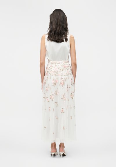 Femme aux cheveux foncés mi-longs, portant un haut blanc sans manches et une longue jupe blanche ornée d'une broderie florale rose, debout de dos.