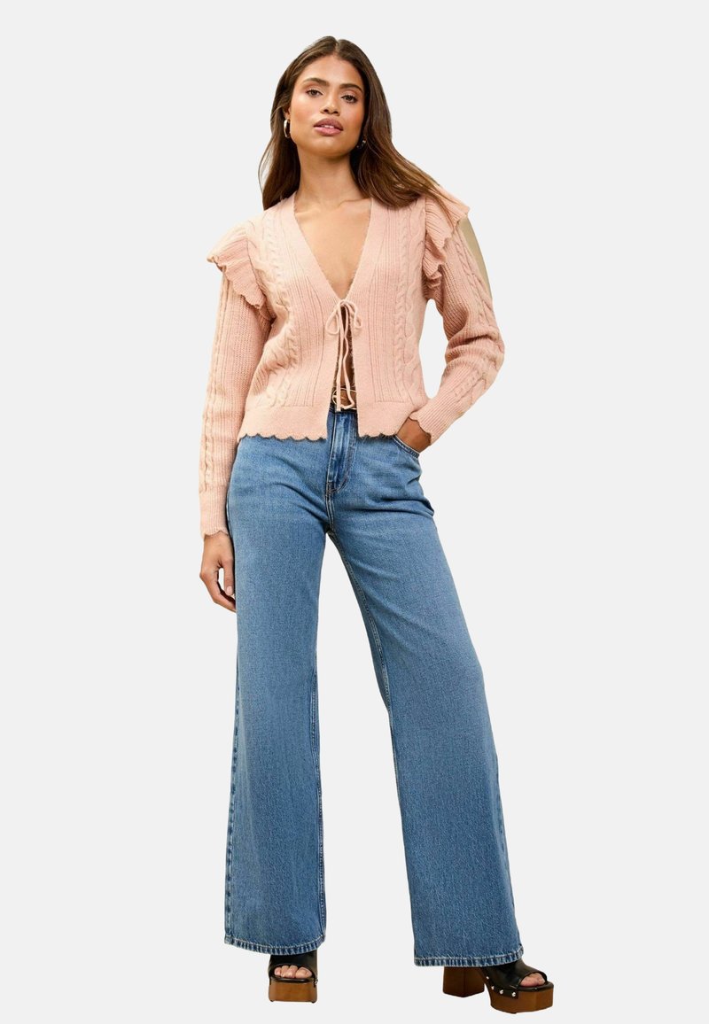 Rožinė megzta kardiganas su raukšlėta tekstūra, giliu V-formos kaklu, priekyje rišamomis detalėmis ir raukšlėtais rankovėmis, derinamas su plačia kojomis mėlynomis džinsinėmis kelnėmis ir platforminėmis bateliais.