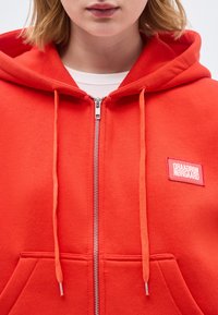 Prim-plan cu o persoană care poartă un hanorac portocaliu aprins cu fermoar și șnururi, având un patch dreptunghiular pe partea stângă a pieptului.