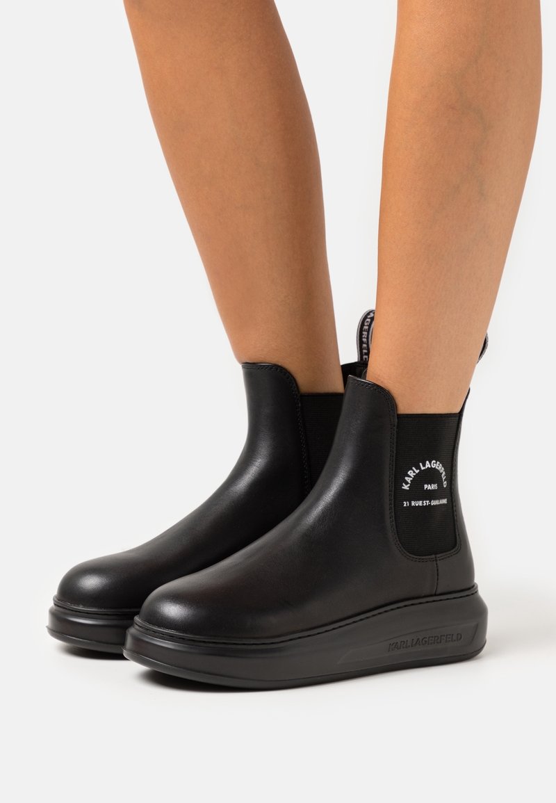 Zwarte enkelboots met dikke zolen gedragen op blote benen, met elastische zijpanelen en witte merktekst op het hielgebied.