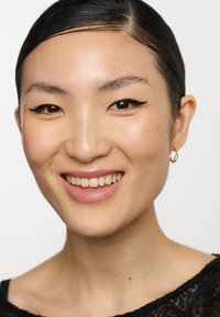 Primo piano di una persona con capelli neri lisci e dritti, con eyeliner scuro e rossetto chiaro. Indossa orecchini a cerchio. Lo sfondo è bianco.