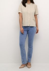 Blusa corto a maniche corte color beige chiaro con colletto arricciato, chiusura con bottoni; indossata con jeans skinny blu chiaro e tacchi nudi.