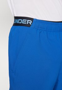Pantalones cortos deportivos azules con una cinturilla elástica negra que presenta "UNDER" en texto azul claro; incluyen bolsillos laterales y una textura de tela suave.
