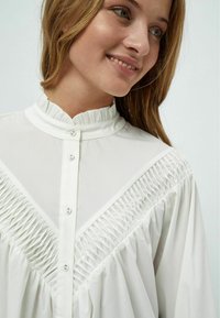 Blusa bianca con colletto alto a balze, chiusura a bottoni e dettagli arricciati lungo il petto e le maniche per un tocco di texture aggiuntivo.