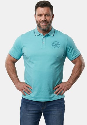 Uomo con la barba che indossa una polo azzurro chiaro e jeans blu, in piedi con le mani sui fianchi, sorridente su sfondo bianco.