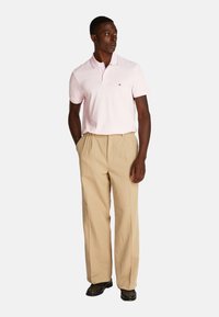 Polo de color rosa claro con collar y mangas cortas, combinado con pantalones de tela color caqui con pliegues. Presenta un logo en la camiseta.