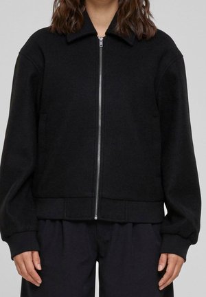 Femme portant une veste noire zippée avec col et manches longues, associée à un pantalon noir ample, debout devant un fond uni.
