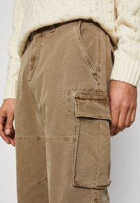 Close-up van een persoon in beige cargobroeken met klepzakken en een crème kabelgebreid sweater, handen ontspannen langs de zijkanten.