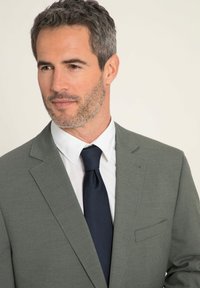 Grauhaar blazer mit Reverskragen und einer einzigen Tasche, kombiniert mit einem weißen Hemd und einer dunkelblauen Krawatte. Glatte Stofftextur; klares Design.