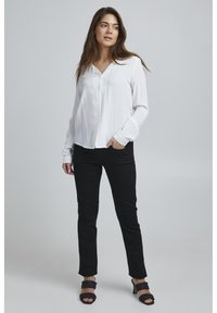 Blusa blanca de manga larga con botones en la parte delantera, combinada con pantalones negros ajustados y sandalias de tacón negro con tiras. Textura suave en toda la prenda.