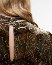 Blouse en velours vert avec des motifs floraux en orange et marron. Elle présente une ouverture en forme de clé au dos et une étiquette en métal doré portant la marque au niveau du cou.