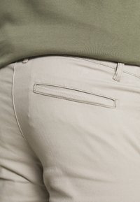 Beige katoenen broek met een achterzak. De stiksels zijn bijpassend en de stof heeft een gladde textuur. De tailleband is eenvoudig.
