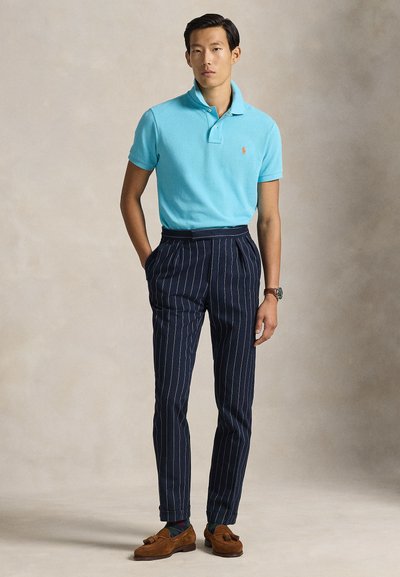 Ανοιχτό μπλε πόλο πουκάμισο, μπλε σκούρο παντελόνι με λεπτές ρίγες, καφέ μοκασίνια. Ρολόι στον αριστερό καρπό, όρθια στάση, ουδέτερο φόντο.