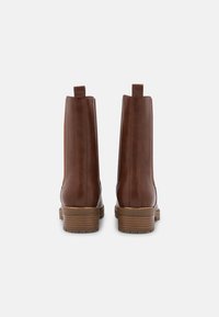 Botines de cuero marrón con textura suave, paneles laterales elásticos y un tacón apilado de madera. Lengüeta en la parte trasera para facilitar el uso.