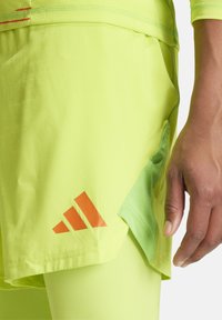 Fluo gele sportshorts met een oranje gestreept logo, voorzien van een gaas zijzak en een gladde stoftextuur, gecombineerd met leggings.