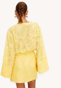 Vestido estructurado amarillo con tela texturizada, mangas largas en forma de campana y cintura ajustada. Presenta un patrón deshilachado para mayor detalle.