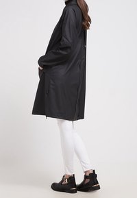 Svart, vattentät lång kappa med hög krage, sidospjäll och avslappnad passform. Kombinerad med vita tajta jeans och svarta ankelboots med utskärningar.