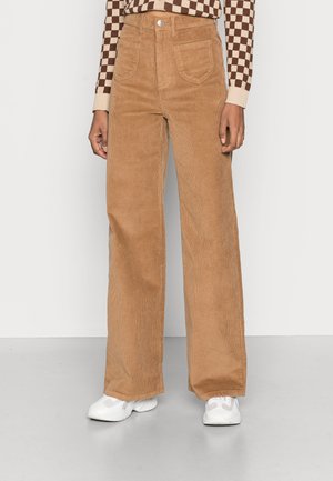 Pantalon large en velours côtelé marron avec un design texturé, taille haute, poches avant et une fermeture lisse à boutons. Porté avec des baskets blanches.