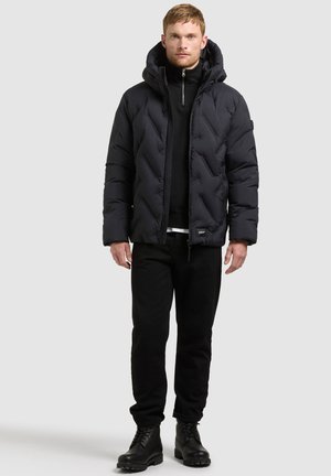 Un homme pose avec une doudoune noire à capuche, un pantalon noir et des bottes à lacets sur un fond clair uni.