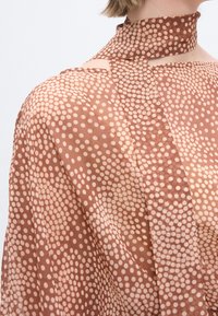 Καφέ ημιδιάφανη μπλούζα με λευκά πουά, λεπτομέρεια με φουλάρι ψηλού λαιμού και ανοιχτόχρωμο κοντό χτένισμα ορατό.