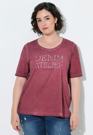 Femme aux cheveux courts et foncés portant des boucles d'oreilles créoles et un t-shirt rouge délavé avec le texte "Denim Atelier est. 1987", associé à un jean bleu.