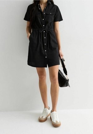 Femme portant une robe courte noire boutonnée avec taille à cordon, baskets blanches, tenant un sac à main noir sur un fond uni.