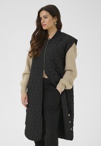 Veste sans manches matelassée noire avec une fermeture éclair à l'avant et des boutons sur les côtés, associée à un pull en maille beige. Présente une coupe longue et fluide.