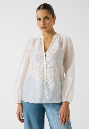 Kobieta z zaczesanymi do tyłu włosami, nosząca złote kolczyki koła, białą zapinaną na guziki bluzkę z kwiatowym haftem i jasnoniebieskie dżinsy na jednolitym tle.