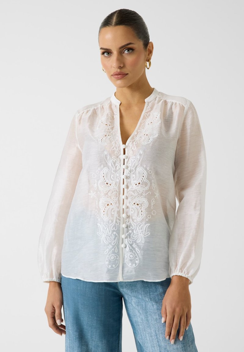 Femme aux cheveux plaqués en arrière, portant des boucles d'oreilles créoles dorées, un chemisier blanc boutonné avec broderie florale et un jean bleu clair sur un fond uni.