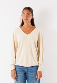 Maglione beige chiaro a V con maniche lunghe, polsini a coste e una vestibilità rilassata. Abbinato a jeans in denim azzurro chiaro. Texture liscia.