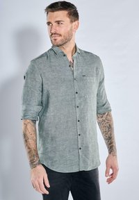 Helles grünes Leinenhemd mit einem Button-Down-Kragen, kurzen hochgerollten Ärmeln, einer Brusttasche und schwarzen Knöpfen, getragen mit dunklen Hosen.
