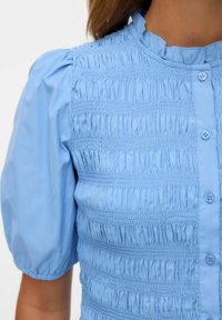 Top azzurro chiaro con texture arricciata, scollo rotondo e maniche a sbuffo. Presenta chiusure con bottoni e un materiale morbido simile al cotone.