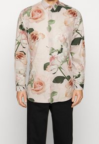 Bloemige shirt met een lichtroze basis en grote rozenpatronen in zachte tinten, lange mouwen, knoopsluiting en een ontspannen pasvorm.