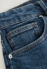 Zili denimi džinsi ar izteiksmīgu aizmugurējo kabatu, kontrastējošām brūnām diega šuvēm un metāla pogas aizdari. Iespējams teksturēts, izbalējis izskats.