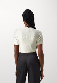 Kortärmad krämfärgad t-shirt med texturerad "PEQUS"-brodyr på ryggen, ihopparad med högmidjade mörkgrå skräddarsydda byxor.