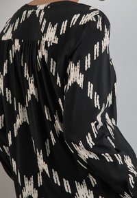 Maglietta nera con motivo astratto crema, caratterizzata da maniche lunghe e un design con schiena arricciata. Tessuto morbido con un aspetto drappeggiato.