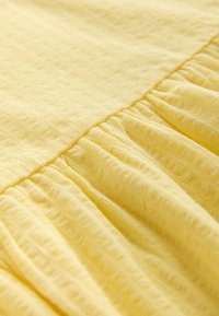 Tissu jaune avec un motif quadrillé texturé et des détails froncés le long d'une couture, créant des plis doux et du volume.