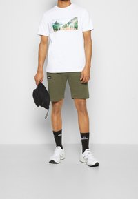 T-shirt bianca in cotone con una stampa grafica colorata, pantaloni corti verde oliva, cappellino nero, calze nere con logo e scarpe da ginnastica bianche.