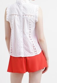 Weiße ärmellose Bluse mit Lochmuster, Rüschenspiegel und Knopfverschluss hinten, kombiniert mit roten Shorts. Glattes, leichtes Material.