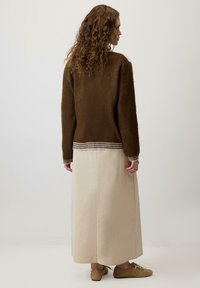 Kobieta z długimi kręconymi włosami, ubrana w brązowy, puszysty sweter, kremową spódnicę do kostek oraz beżowe buty, stojąca na jednolitym białym tle.