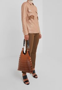 Persoon in een lichtbruine leren jasje met franjes, bruine broek en zwarte open-toe sandalen, met een grote bruine gevlochten handtas.