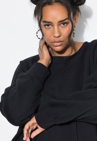 Sudadera negra con cuello redondo, mangas largas y puños acanalados. La tela parece suave y texturizada, acentuada por un brillo sutil.