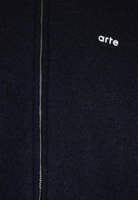 Tessuto scuro con una cerniera verticale argento e il logo "arte" in minuscolo ricamato sul lato superiore destro.