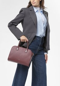 Borsa a mano in pelle marrone con forma strutturata, due manici superiori e una finitura leggermente testurizzata, abbinata a un blazer grigio e una camicia a righe blu.