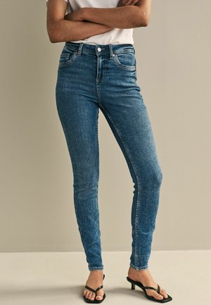 Femme portant un jean skinny bleu taille haute et des sandales noires à petit talon, debout les bras croisés devant un fond uni.