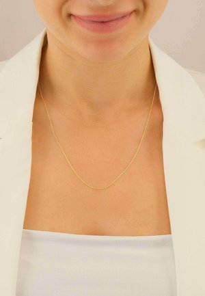 Primo piano di una persona che indossa una sottile collana d'oro con una maglietta bianca e un blazer chiaro.