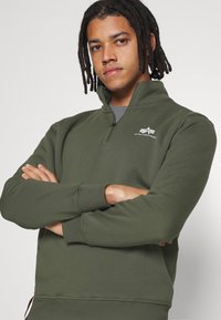 Felpa a collo alto in verde scuro, con zip a un quarto, polsini a costine e logo sul petto. Tessuto morbido con una texture liscia.