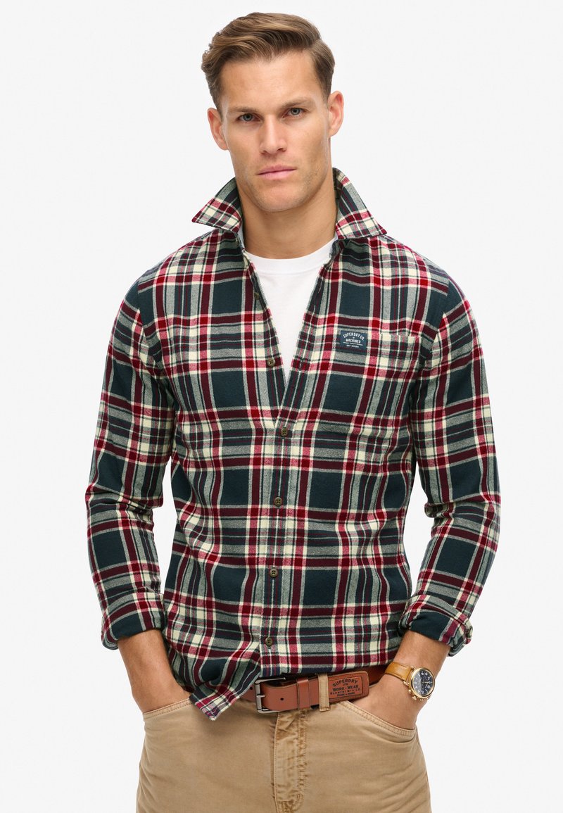 Geruite flanellen overhemd in donker groen, rood en crème; met een knoopsluiting en een logo-patch op de borst. Draagt over een wit T-shirt.